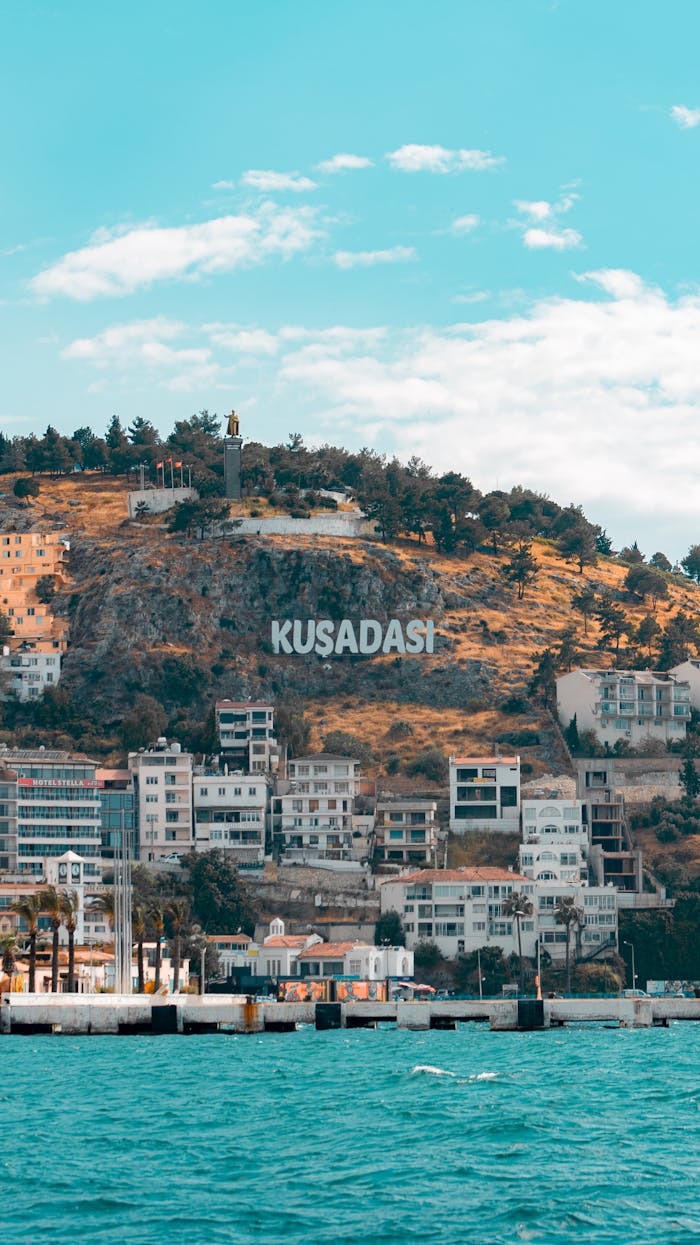 Scenic view of Kusadasi's coastline with hillside landmarks and azure sea under bright sky.